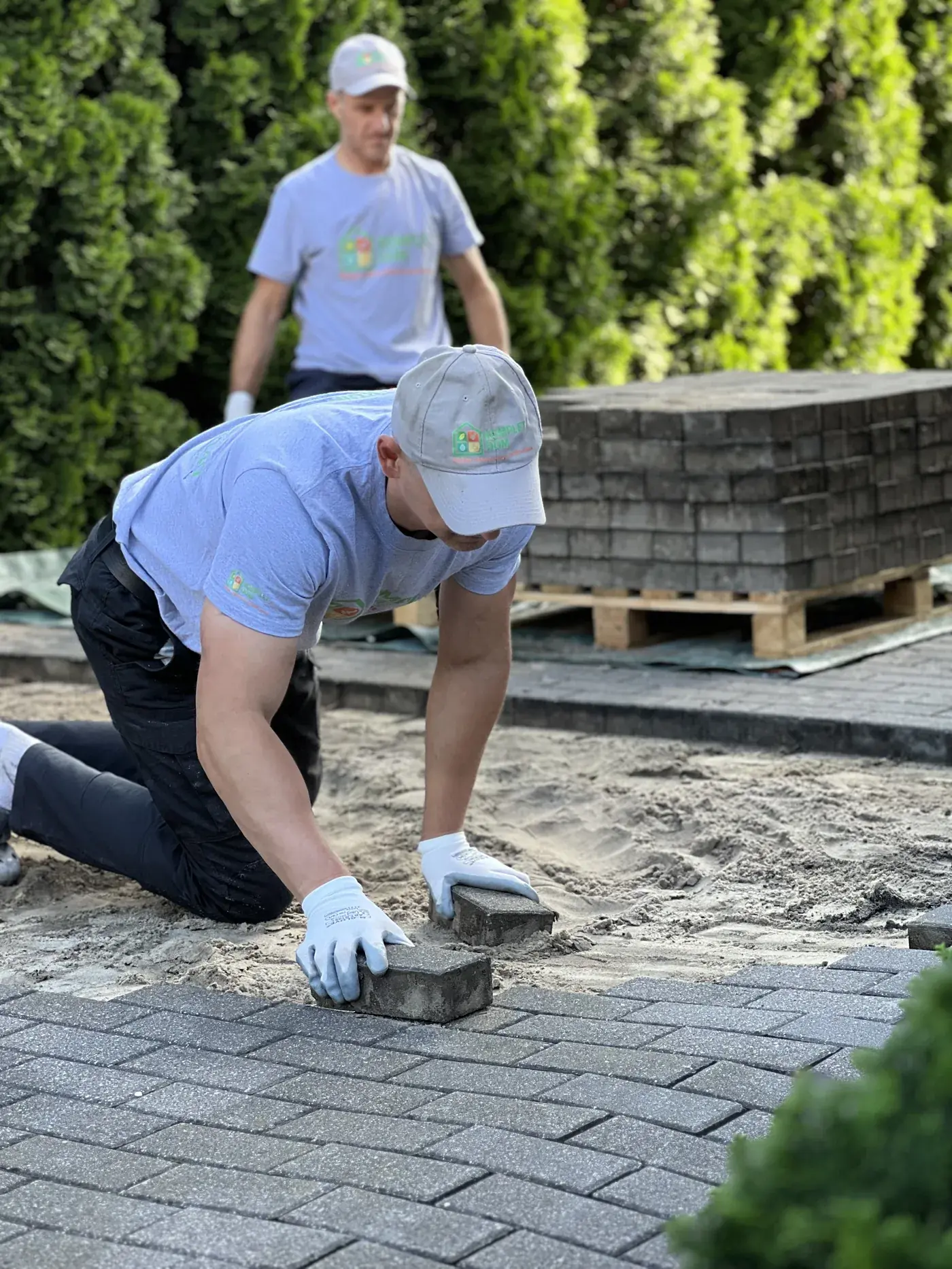 Dwóch pracowników KompletDom w firmowych koszulkach układa szarą kostkę brukową na podsypce piaskowej
